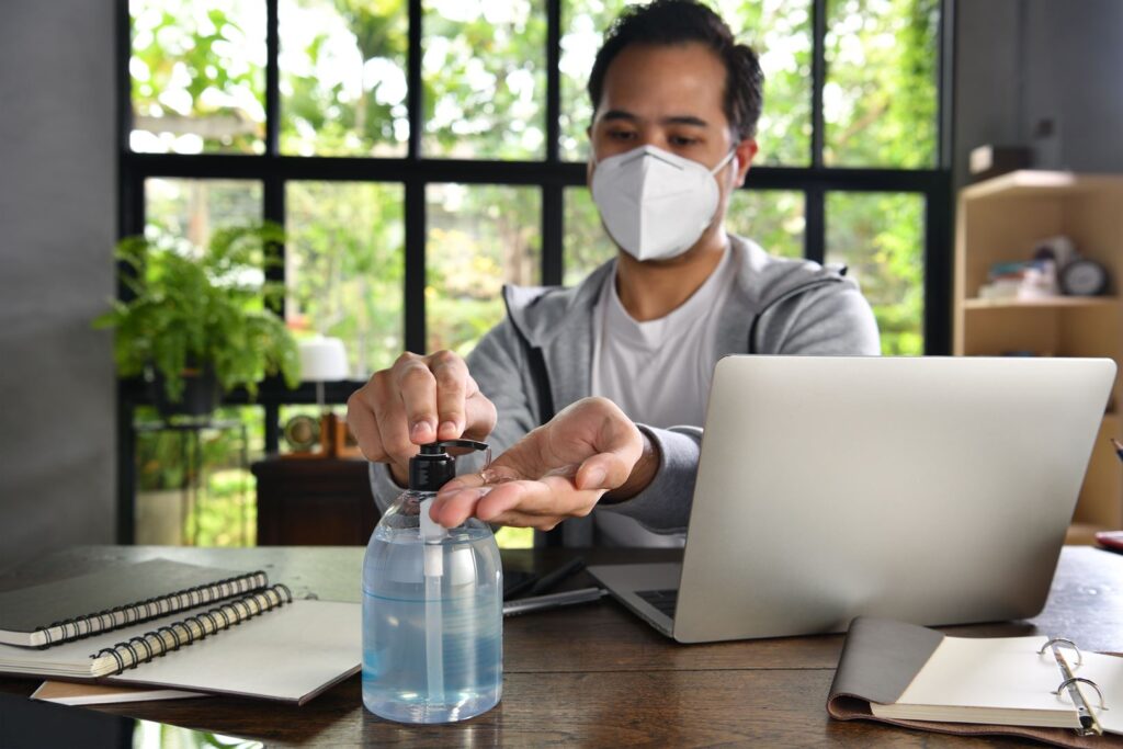 Asian man in quarantine and social distancing wearing surgical mask and cleaning hands with alcohol gel sanitizer while working from home during Covid-19 Coronavirus pandemic